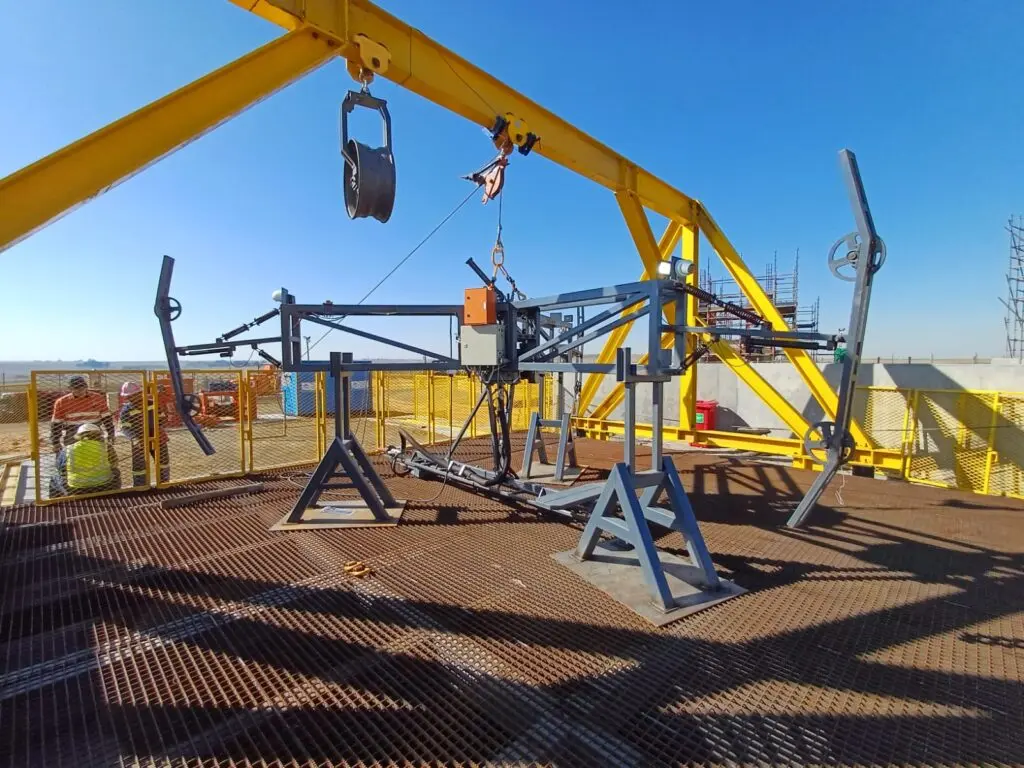 Um equipamento de teste de metal em uma plataforma gradeada sob um céu azul brilhante. A configuração inclui um guindaste de pórtico amarelo e vários componentes metálicos e fios. Materiais de construção e trabalhadores são visíveis ao fundo.