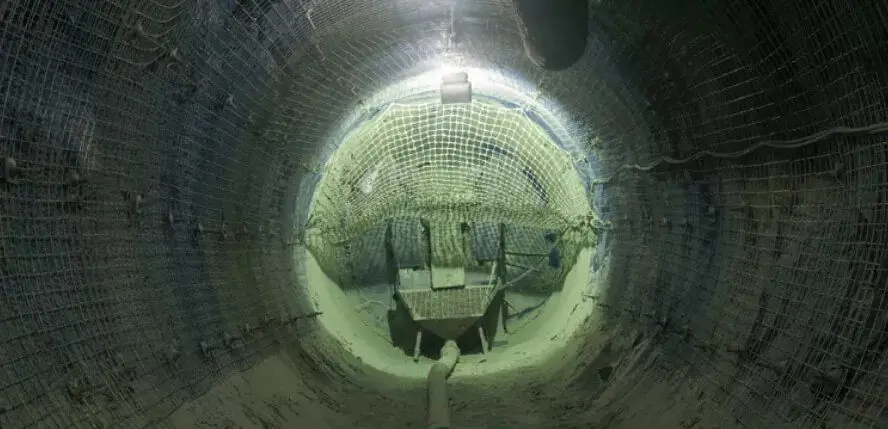 Um túnel subterrâneo mal iluminado, reforçado com redes de metal nas paredes e no teto. O piso é áspero e parece ter equipamentos de construção e detritos espalhados, o que sugere um projeto de escavação em andamento ou concluído recentemente.