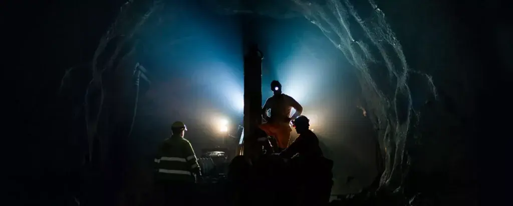 Três mineiros com equipamentos de segurança trabalham no subsolo em um túnel pouco iluminado. Um mineiro está de pé e opera um maquinário iluminado por luzes azuis, enquanto outros dois observam. As paredes rochosas do túnel envolvem a cena, aumentando a sensação de profundidade e espaço fechado.
