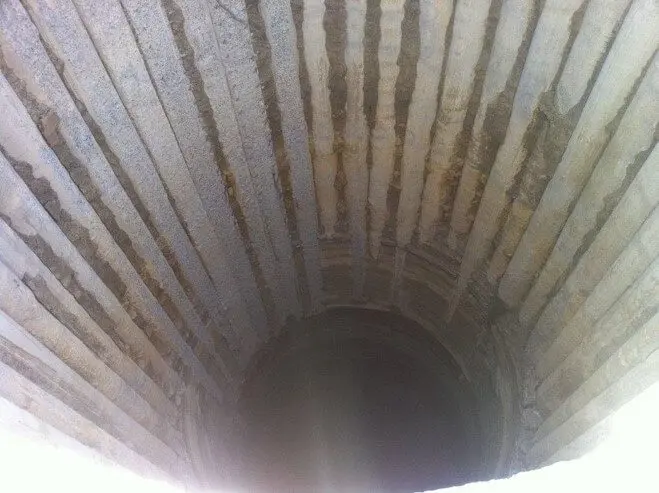Um poço profundo e circular com paredes de pedra caneladas que descem para a escuridão. A luz na abertura lança sombras, enfatizando a superfície texturizada e a profundidade do poço.