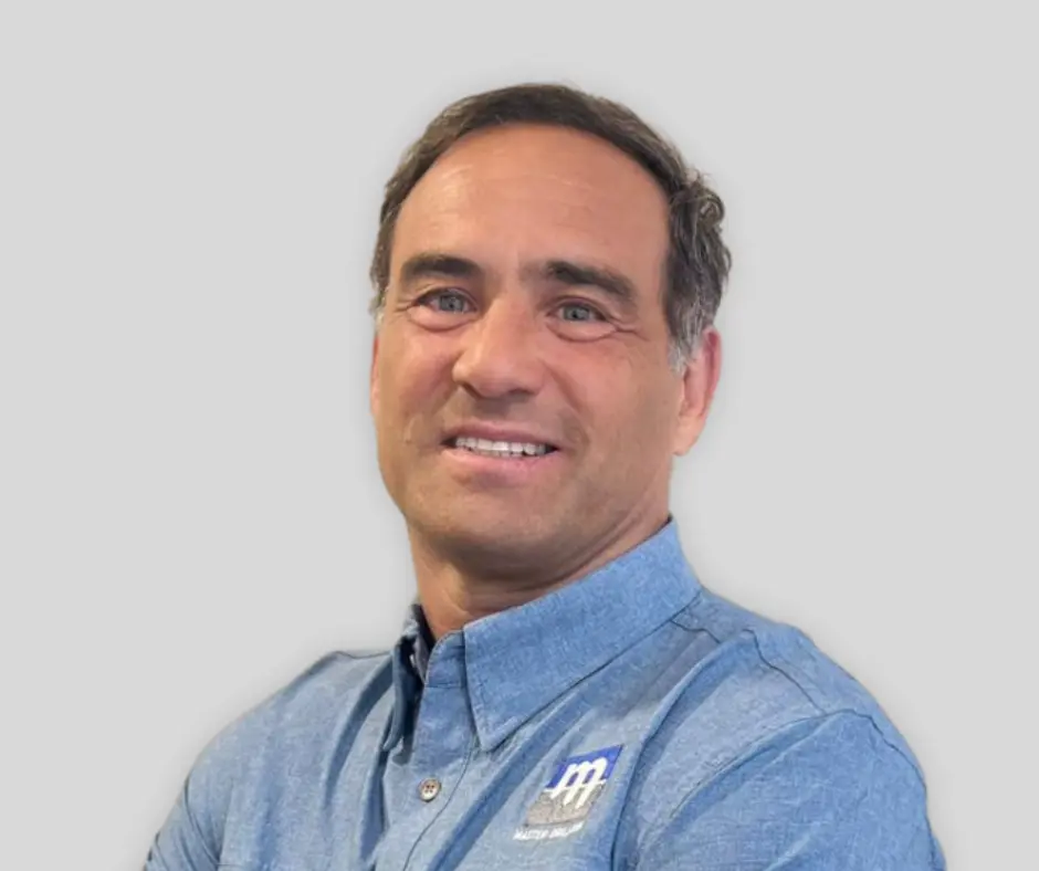A man with short brown hair wearing a blue button-up shirt with a white and blue logo on the chest, smiling in front of a plain light gray background.