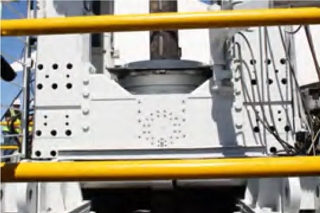 Close-up of industrial machinery with a white metal structure, various bolts, and black cables, framed by yellow safety rails. The machinery appears to be part of a larger mechanical setup, possibly in a factory or construction site.