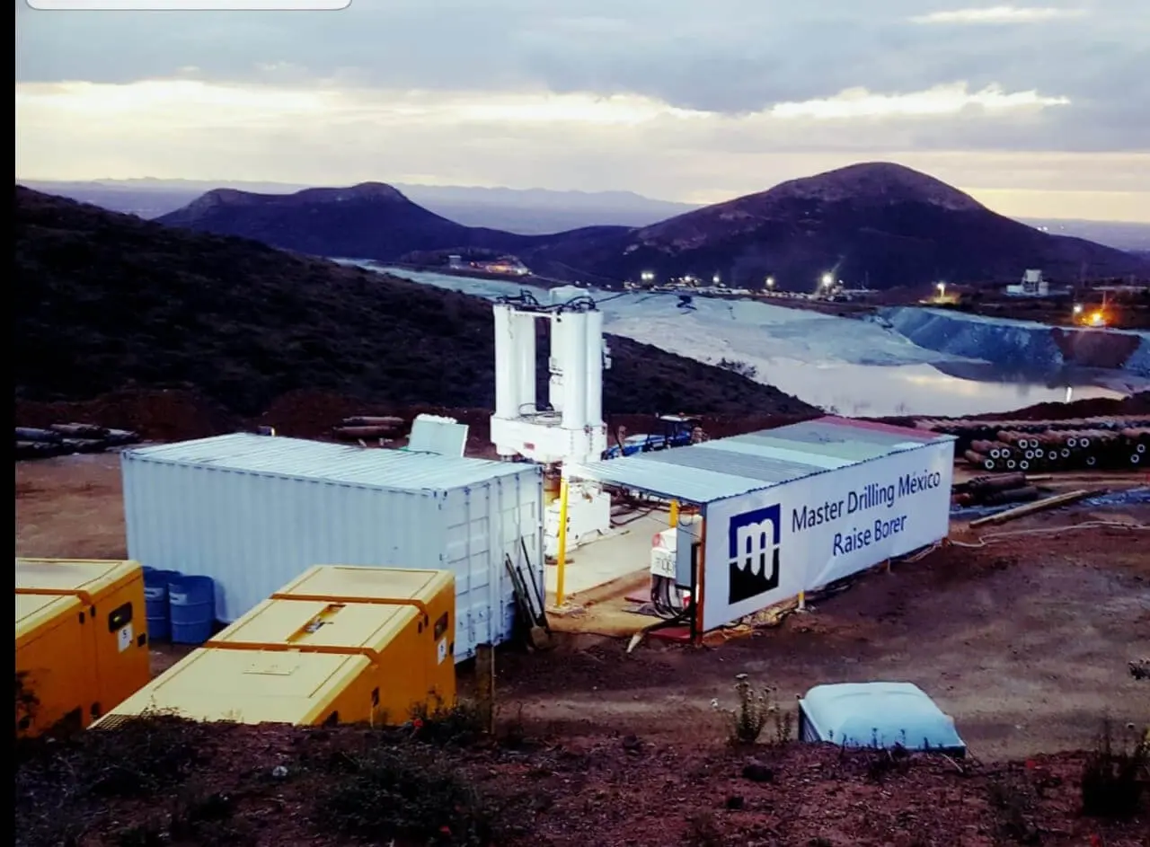 A Master Drilling México raise borer site at dusk, with white containers, yellow generators and floodlit mine workings among mountains.