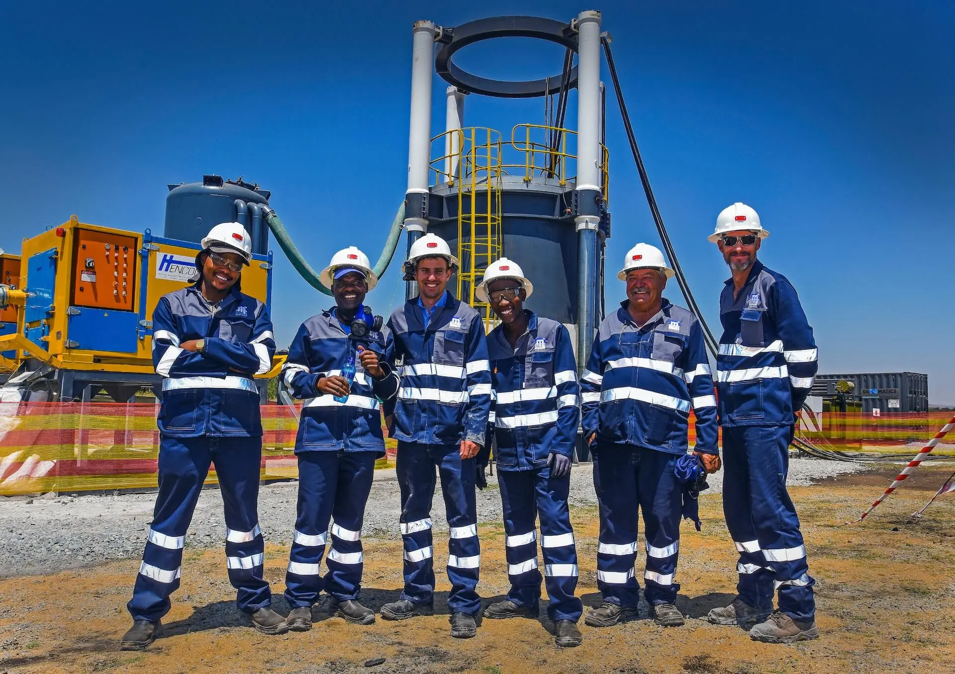 Six crew members in navy overalls and white hard hats stand in front of a raise bore rig and support equipment on an open drilling site.