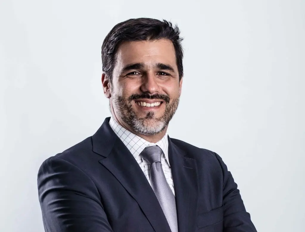 A man in a dark suit, light shirt, and tie stands smiling with arms crossed against a plain light background. He has short dark hair, a beard, and mustache.