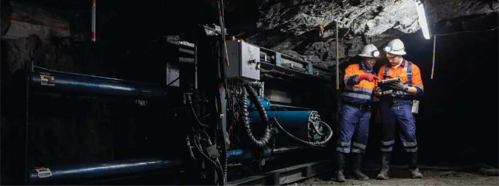 Dois mineiros usando capacetes e equipamentos de segurança estão em um túnel subterrâneo escuro iluminado por uma única luz. Eles estão trabalhando com maquinário e parecem estar coordenando ou verificando algo em um tablet. O plano de fundo é robusto, com rochas e equipamentos de mineração.