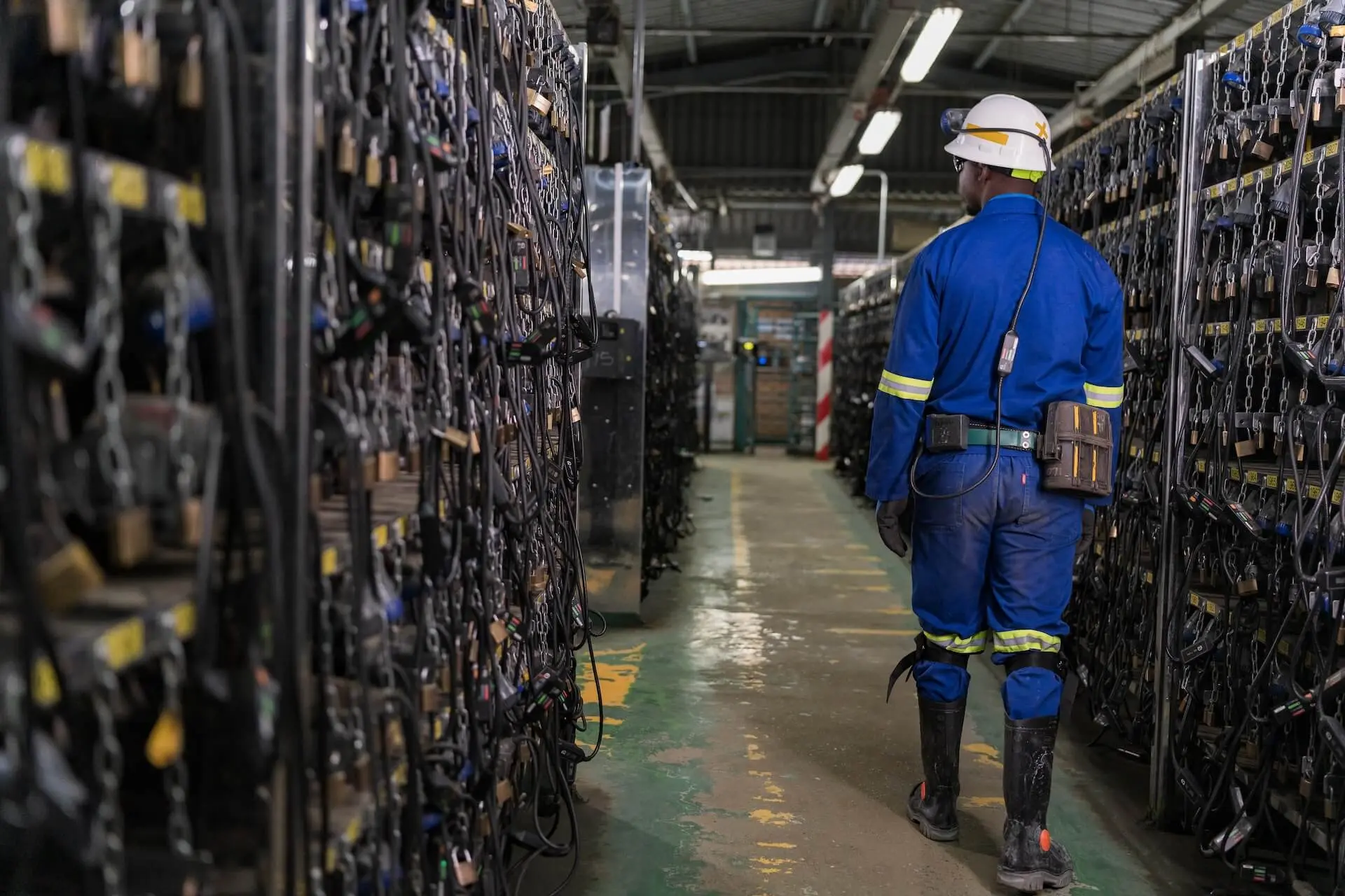 Um trabalhador com roupa de proteção azul caminha por uma sala de lâmpadas de mina ladeada por equipamentos de carregamento.