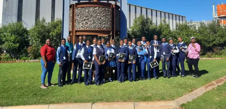 A large group of students in matching blue school uniforms is posing for a photo in front of an industrial structure with a large "M" logo. Two adults in casual clothing are standing on either side of the group. The sky is clear and blue.