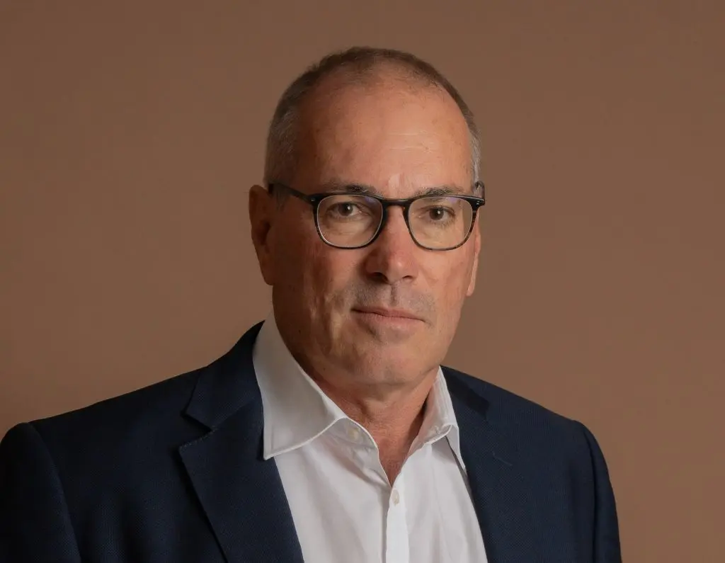 A middle-aged man wearing glasses, a white shirt, and a dark blazer poses against a plain, light brown background. He has short hair and a neutral expression.