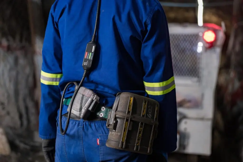 Um trabalhador vestindo roupas de proteção azuis com faixas amarelas refletivas e equipamento preso ao cinto é mostrado por trás. O trabalhador está dentro do que parece ser uma caverna ou um túnel subterrâneo, com maquinário e pouca iluminação ao fundo.
