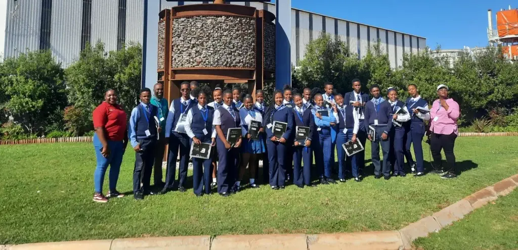 Students in blue school uniforms gather with adults in front of a Master Drilling facility.