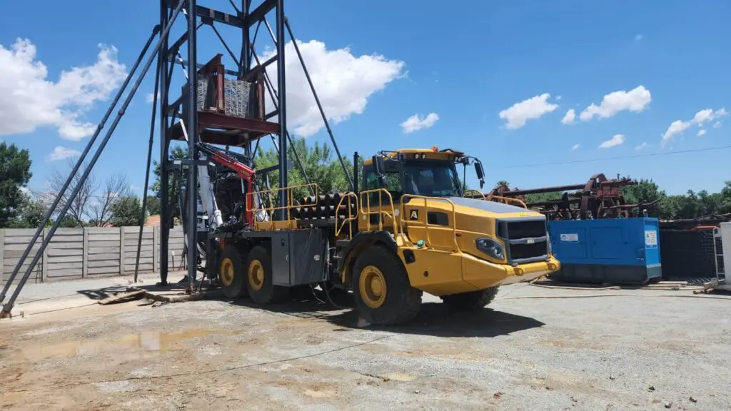 Um grande veículo de construção amarelo está posicionado próximo a uma estrutura metálica alta em uma área de trabalho externa. O céu azul claro está pontilhado com algumas nuvens, e vários equipamentos e materiais estão espalhados pelo local.