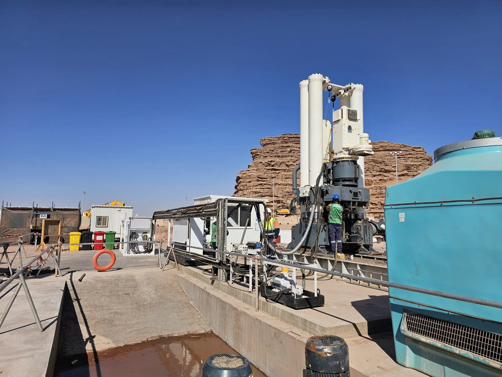 A raise bore rig with tall white cylinders on a concrete civil site, with workers in safety gear and desert rock formations behind.
