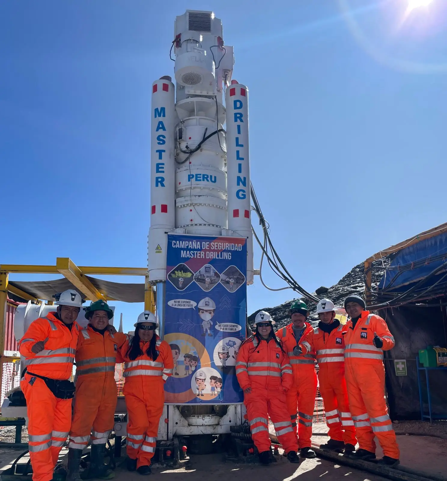 Seven crew members in orange overalls give thumbs up beside a white Master Drilling Peru raise borer carrying a safety-campaign banner.
