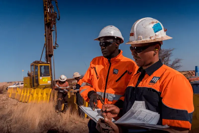 Dois trabalhadores vestindo equipamentos de segurança laranja e azul e capacetes brancos discutem algo enquanto seguram documentos em um local de mineração ou construção sob um céu azul claro. Uma plataforma de perfuração é visível ao fundo, ao lado de outros trabalhadores envolvidos em tarefas.