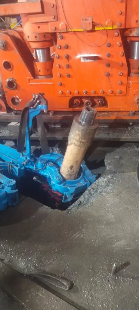 Um braço mecânico segura um objeto cilíndrico, em frente a uma máquina industrial laranja. A área ao redor da máquina parece ser um chão de oficina empoeirado ou sujo. A cena provavelmente retrata parte de um processo de fabricação ou reparo.