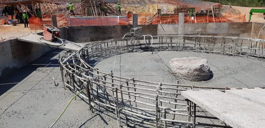 Local de construção mostrando uma estrutura circular de concreto com vergalhões de aço e concreto parcialmente derramado. Trabalhadores com equipamentos de segurança estão próximos, com redes de proteção laranja e equipamentos ao redor da área. Uma grande rocha é visível dentro da estrutura.