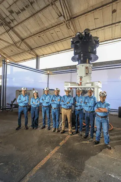 Um grupo de oito pessoas com uniformes azuis e capacetes de proteção está dentro de um grande armazém industrial. Elas estão em frente a um equipamento industrial considerável, com uma iluminação significativa vindo de trás. O armazém tem um teto alto e arqueado.