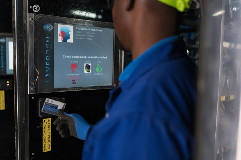 Pessoa vestindo roupa de proteção azul e capacete amarelo neon verificando o status do equipamento em uma tela usando um cartão. A tela exibe gráficos e texto para checkout de equipamentos com indicadores destacando 'validação falhou' em vermelho.