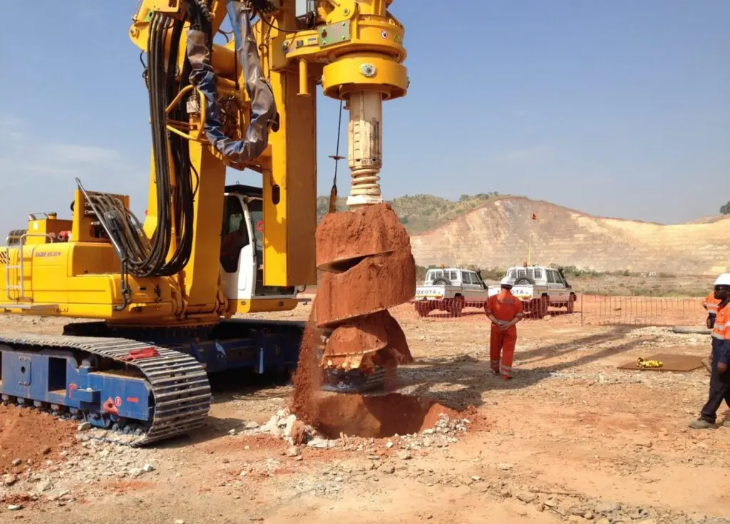 Uma grande máquina de perfuração amarela com um acessório de trado em espiral está em operação em um canteiro de obras. Trabalhadores com equipamento de segurança laranja estão próximos, supervisionando o trabalho. O plano de fundo apresenta uma paisagem montanhosa e vários veículos utilitários.